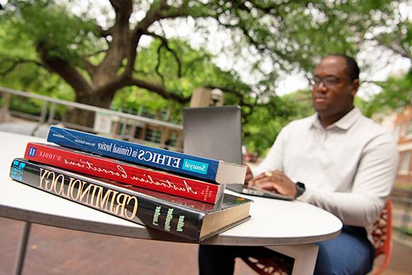 Close up of criminal justice books on an outdoor table with a student typing on his laptop in the backg