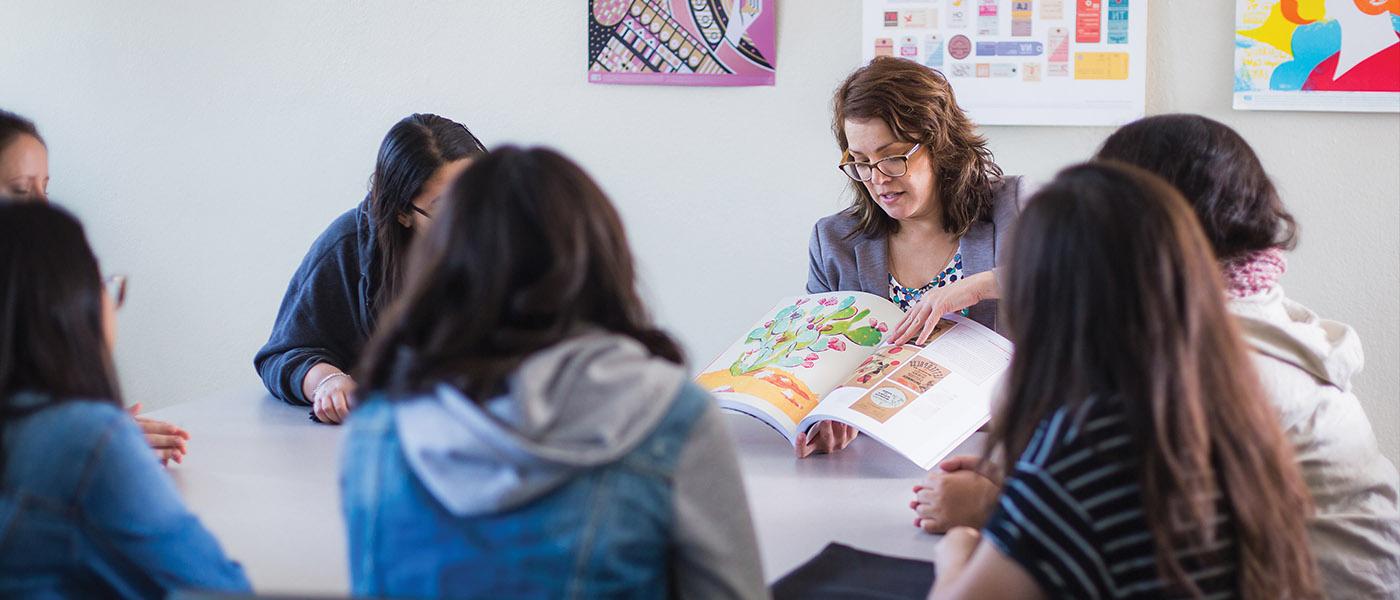 group of graphic design students listening to their professor as she shows them work in a design book